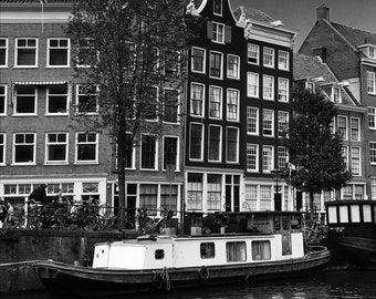 May include: A black and white photo of a canal in Amsterdam, Netherlands. A small boat is docked in the canal. The boat is white with a black bottom. The boat is in front of a row of historic buildings with black and white windows. The buildings are lined up along the canal.