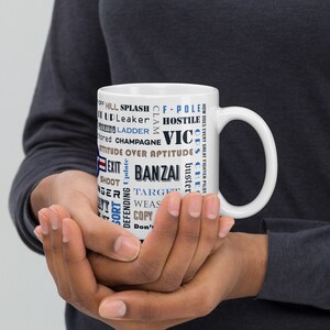 Pode incluir: Caneca de cerâmica branca com um design de nuvem de palavras azul e marrom. As palavras incluem "banzai", "hostile", "attitude", "exit", "vic", "target", "shoot", "ladders", "champagne", "leaker", "splash", "f-pole", "update", "wea", "copy", "don't", "buster", "che", "off", "hill", "claim", "how does every great fighter pilot", "and more".