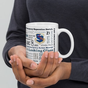 Pode incluir: Caneca de cerâmica branca com um design preto e azul que inclui as palavras "Emergency Separation Switch", "Laydown Bombs", "DEFCON", "HHCL H-Hour Control Line", "LADDS", "elobber Factor", "Emergency Action Message", "Looking Glass", "Combat Mission Folder", "Alternate Landing Sites", "MIL-National Dome", "Dead" e "De". O design também inclui o emblema da Força Aérea.