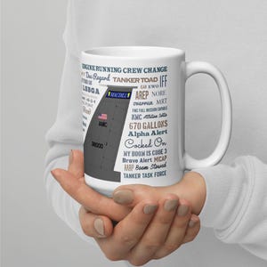 Może przedstawiać: Biały ceramiczny kubek z grafiką samolotu-cysterny, tekstem "MACDILL" i "AMC". Dodatkowy tekst zawiera "670 GALLONS", "Alpha Alert" i "Tanker Task Force". Kubek jest trzymany przez osobę.