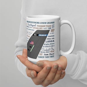 Może przedstawiać: Biały ceramiczny kubek z grafiką samolotu, napisem "TRAVIS" i amerykańską flagą. Kubek jest ozdobiony terminami lotniczymi, takimi jak "ENGINE RUNNING CREW CHANGE" i "TANKER TASK FORCE".