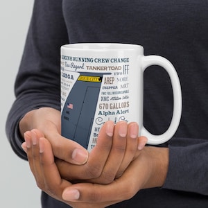 Może przedstawiać: Biały ceramiczny kubek z niebiesko-szarą grafiką samolotu i tekstem, w tym "SIOUX CITY", "670 GALLONS" i "Alpha Alert". Kubek ma duży uchwyt i jest trzymany przez osobę.