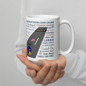 Może przedstawiać: Biały ceramiczny kubek z graficznym wzorem. Projekt zawiera tekst, taki jak "ENGINE RUNNING CREW CHANGE" i "TANKER TOAD". Projekt zawiera stylizowany element samolotu z amerykańską flagą i słowem "MCGUIRE".