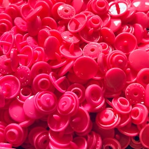 May include: A large pile of bright pink plastic snap buttons. The buttons are all the same size and shape, and they have a small hole in the centre.