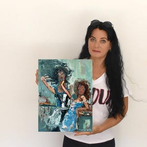 May include: An abstract painting featuring two women in blue dresses. The artwork has a textured, brushstroke style with a palette of blues, whites, and browns. The painting is held by a person in a white t-shirt.