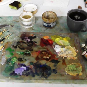 May include: An artist's workspace with a palette of mixed paint colors, brushes, and paint tubes. A gray mug of coffee sits next to the palette. Jars of paint and a palette knife are also visible, suggesting a creative environment.