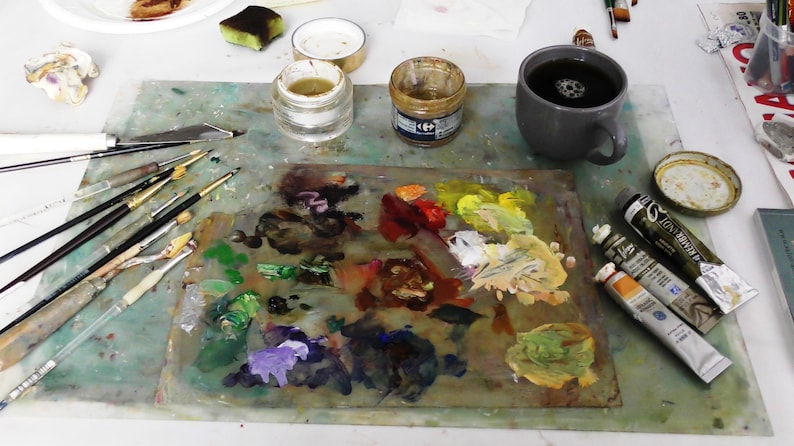 May include: An artist's workspace with a palette of mixed paint colors, brushes, and paint tubes. A gray mug of coffee sits next to the palette. Jars of paint and a palette knife are also visible, suggesting a creative environment.