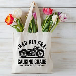 K&ouml;nnte beinhalten: Eine wei&szlig;e Canvas-Tragetasche mit schwarzem Text und einer Motorradgrafik. Der Text lautet "Rad Kid Era Causing Chaos - Life in the Fast Lane".