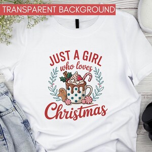 Peut inclure: T-shirt blanc avec un motif de No&euml;l. Le design comprend le texte "JUST A GIRL who loves Christmas" en rouge, une tasse de chocolat chaud, un bonhomme en pain d'&eacute;pices et des cannes en sucre.