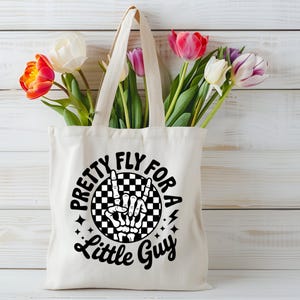 Puede incluir: Un bolso tote de color crema con el texto "Pretty Fly For A Little Guy" en negro. El dise&ntilde;o incluye una mano de esqueleto haciendo un signo de la paz dentro de un c&iacute;rculo a cuadros en blanco y negro. La bolsa est&aacute; llena de tulipanes coloridos.