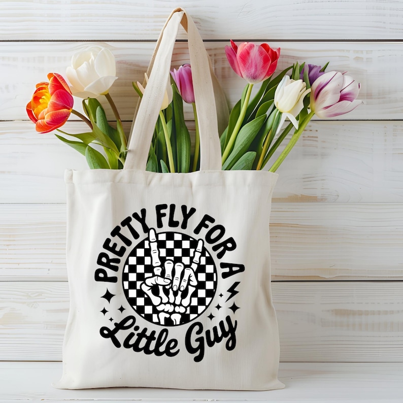 Puede incluir: Un bolso tote de color crema con el texto "Pretty Fly For A Little Guy" en negro. El dise&ntilde;o incluye una mano de esqueleto haciendo un signo de la paz dentro de un c&iacute;rculo a cuadros en blanco y negro. La bolsa est&aacute; llena de tulipanes coloridos.