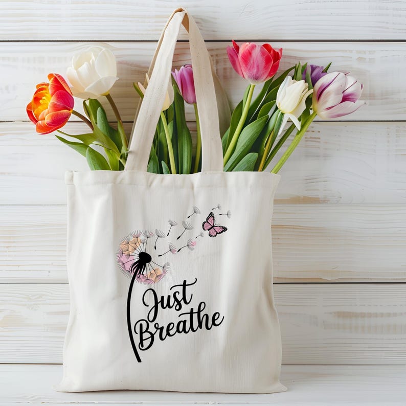 Może przedstawiać: Kremowa torba z nadrukiem dmuchawca i napisem "Just Breathe" czarną czcionką. Dmuchawiec ma r&oacute;żowe i brzoskwiniowe akcenty, z r&oacute;żowym motylem. Torba jest wypełniona kolorowymi tulipanami.