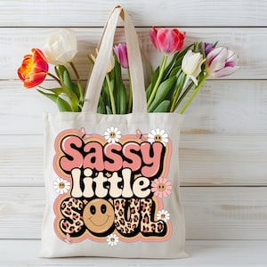 Puede incluir: Una bolsa de tela blanca con un gr&aacute;fico rosa y naranja que dice "Sassy little soul" con una carita sonriente y estampado de leopardo. El gr&aacute;fico est&aacute; rodeado de margaritas blancas y mariposas.