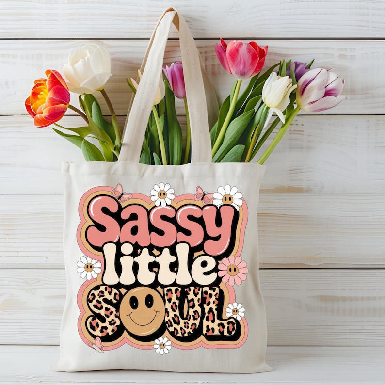 Puede incluir: Una bolsa de tela blanca con un gr&aacute;fico rosa y naranja que dice "Sassy little soul" con una carita sonriente y estampado de leopardo. El gr&aacute;fico est&aacute; rodeado de margaritas blancas y mariposas.