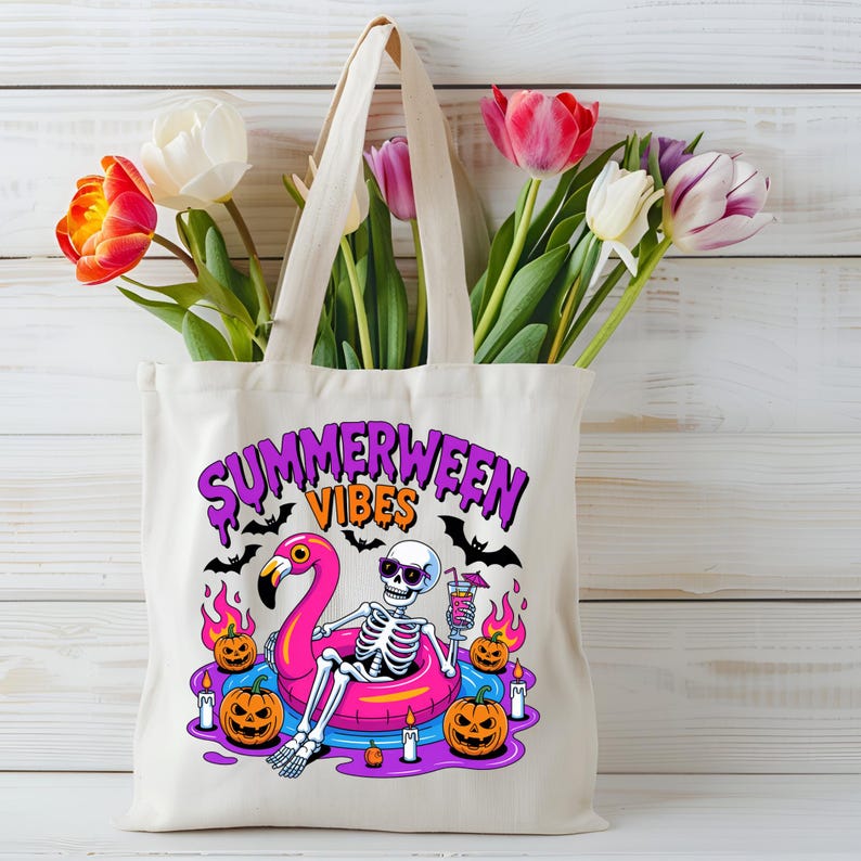 May include: Cream-colored tote bag with the words "SUMMERWEEN VIBES" in purple and orange. The graphic features a skeleton wearing sunglasses, floating on a pink flamingo pool float, with pumpkins and bats. The bag is filled with colorful tulips.