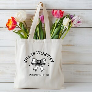 Op de afbeelding: Een witte canvas tote bag met zwarte tekst die "She is Worthy" zegt met een strikgrafiek en "Proverbs 31" eronder. De tas is gevuld met een boeket roze, witte en oranje tulpen.