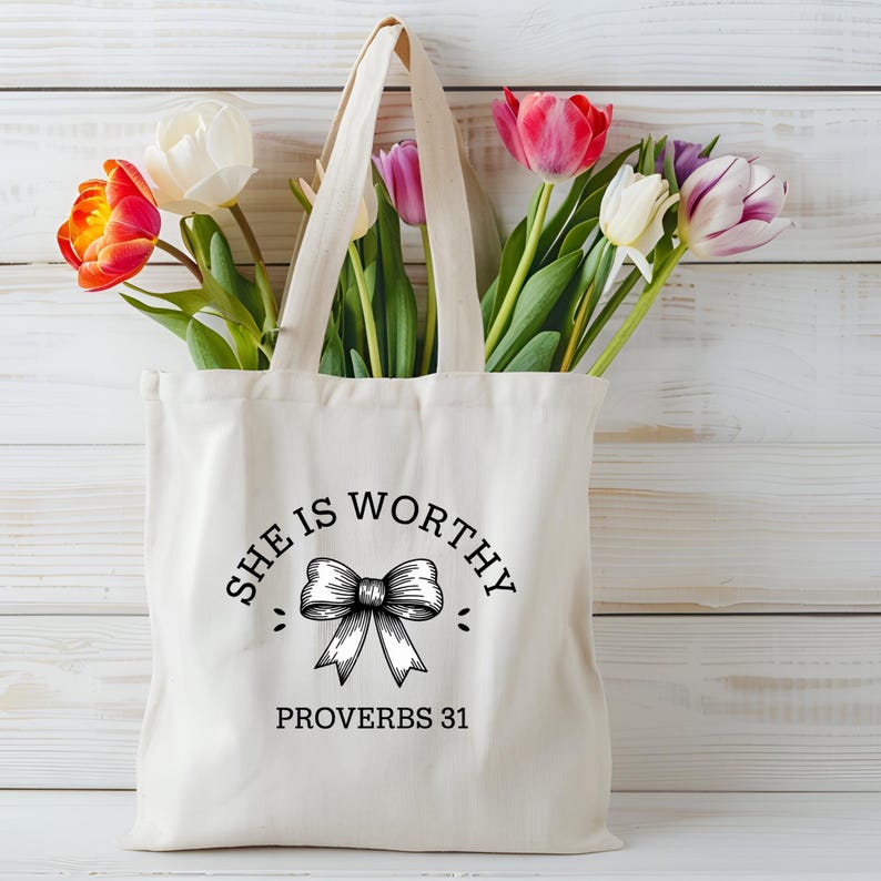 Op de afbeelding: Een witte canvas tote bag met zwarte tekst die "She is Worthy" zegt met een strikgrafiek en "Proverbs 31" eronder. De tas is gevuld met een boeket roze, witte en oranje tulpen.
