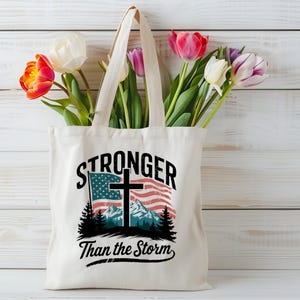 May include: A white canvas tote bag with a graphic design featuring a cross over an American flag with the text "Stronger Than the Storm".