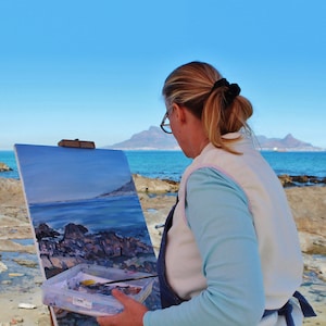 May include: A woman paints a seascape on a canvas easel on a rocky beach. The painting depicts a blue ocean with rocky outcroppings and a distant mountain range. The woman is wearing a white vest over a blue shirt and has her hair pulled back in a ponytail.