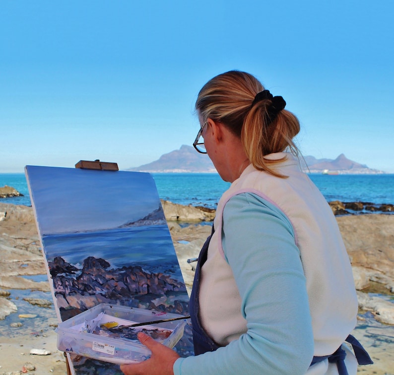May include: A woman paints a seascape on a canvas easel on a rocky beach. The painting depicts a blue ocean with rocky outcroppings and a distant mountain range. The woman is wearing a white vest over a blue shirt and has her hair pulled back in a ponytail.