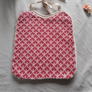 Peut inclure: Une bavette à motif rose et or avec une bordure blanche et une cravate blanche. La bavette a un motif géométrique de triangles qui se chevauchent.