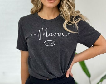 May include: A dark gray t-shirt with the word "Mama" in white cursive script and a small heart on either side. Below the word "Mama" is a white oval with the text "Est. 2023" inside.