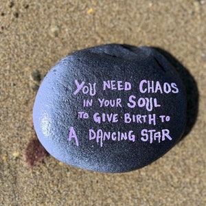 May include: A dark gray painted rock with the words "You need chaos in your soul to give birth to a dancing star" in lavender script. The rock is resting on a sandy surface.