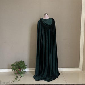 May include: A dark green hooded cloak made of velvet fabric. The cloak is displayed on a mannequin, showcasing its length and design. The hood is visible, and the cloak drapes to the floor. The background is a neutral wall with a plant in the corner.