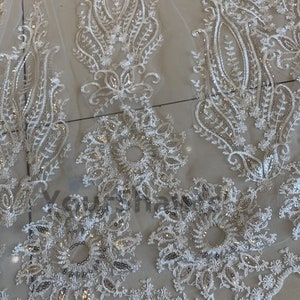 May include: White lace fabric with a delicate floral pattern and silver sequins. The fabric has a scalloped edge.
