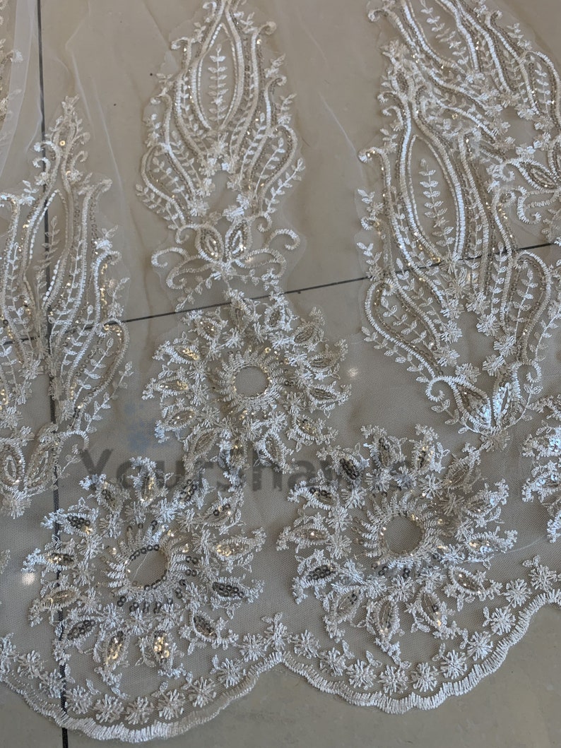 May include: White lace fabric with a delicate floral pattern and silver sequins. The fabric has a scalloped edge.