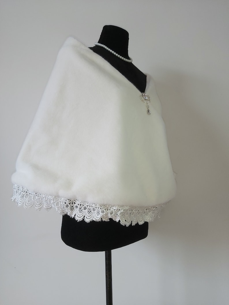 May include: White faux fur shawl with a lace trim. The shawl is displayed on a black mannequin.