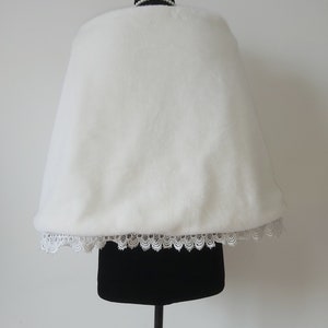 May include: White faux fur bridal shawl with a lace trim. The shawl is displayed on a black mannequin.