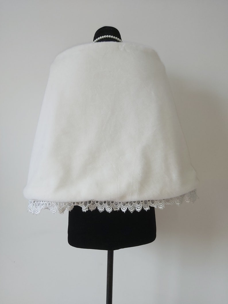 May include: White faux fur bridal shawl with a lace trim. The shawl is displayed on a black mannequin.
