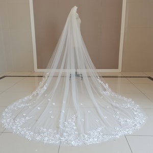May include: A long white tulle wedding veil with a floral lace trim. The veil is displayed on a mannequin in a white room.