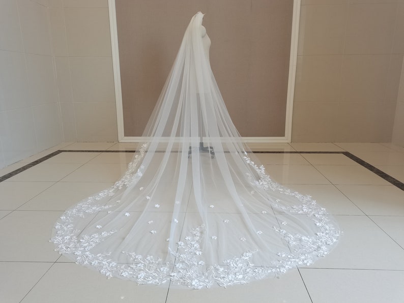 May include: A long white tulle wedding veil with a floral lace trim. The veil is displayed on a mannequin in a white room.