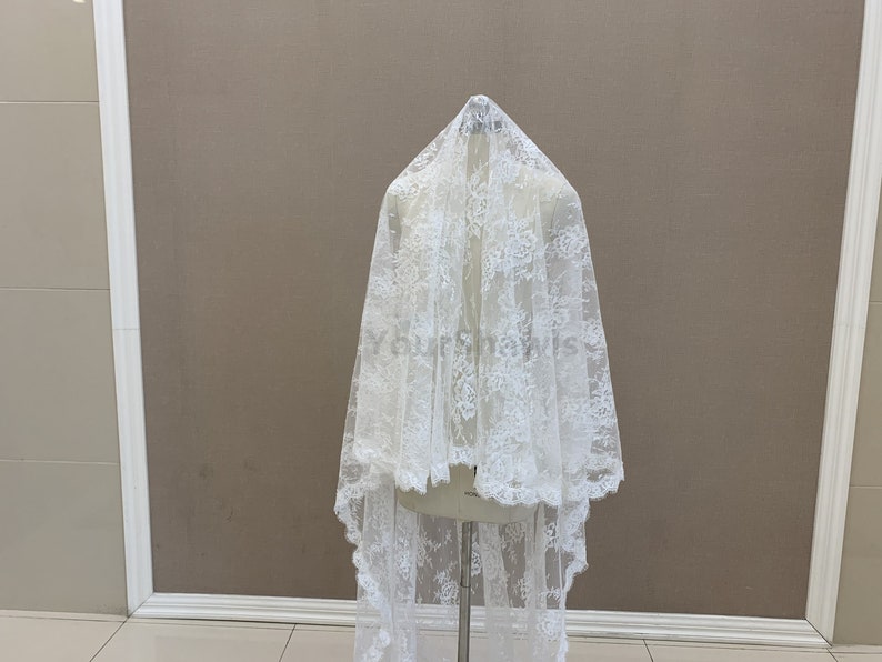 May include: White lace fabric with a delicate floral pattern. The fabric is draped over a mannequin and has a scalloped edge.