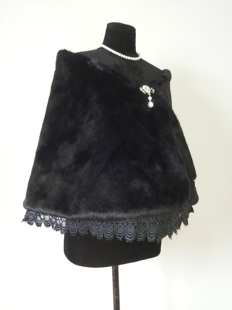 May include: A black faux fur shawl with a black lace trim. The shawl is draped over a black mannequin and has a silver brooch with a dangling crystal.