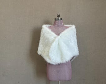 Może przedstawiać: Biały szal ze sztucznego futra narzucony na manekina. Szal ma miękką, puszystą fakturę i zaokrąglony kształt, przeznaczony do noszenia na ramionach. Manekin ma stonowany różowy kolor, na neutralnym tle.