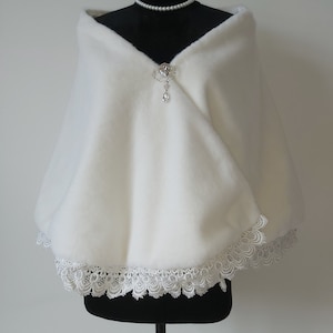May include: White faux fur shawl with a lace trim and a crystal brooch. The shawl is draped over a black dress form.