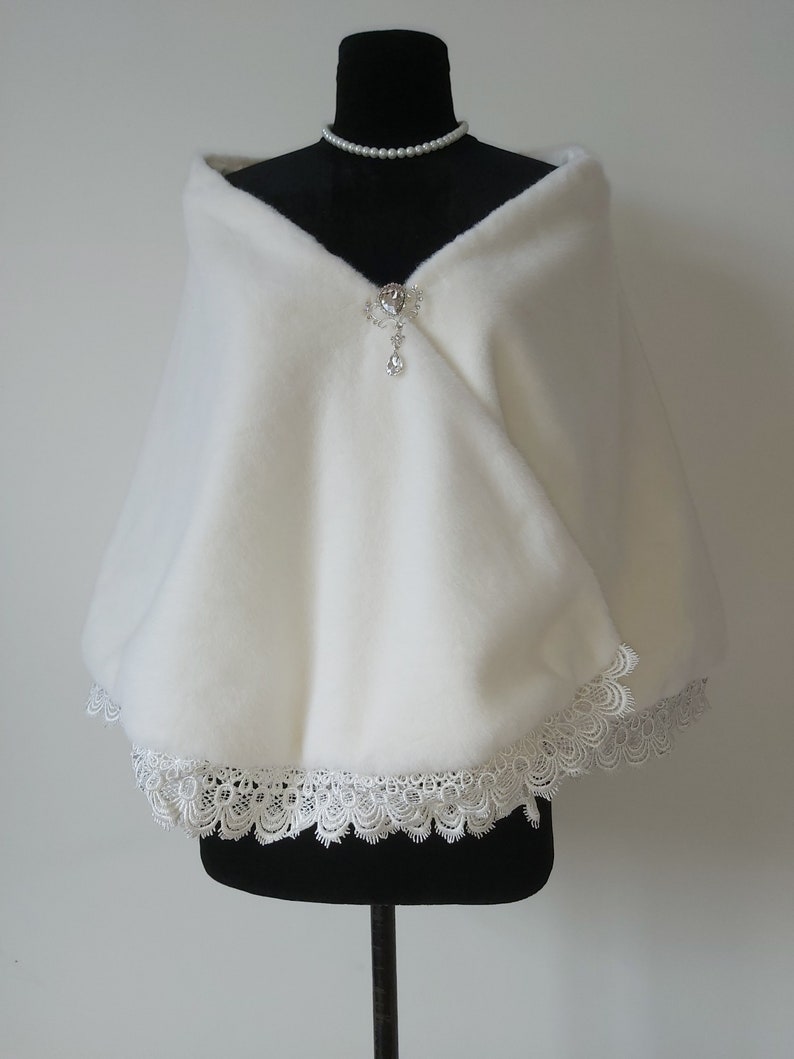May include: White faux fur shawl with a lace trim and a crystal brooch. The shawl is draped over a black dress form.