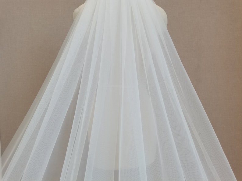 May include: A white tulle wedding veil with a single layer of soft, sheer fabric. The veil is draped over a dress form and has a flowing, elegant design.