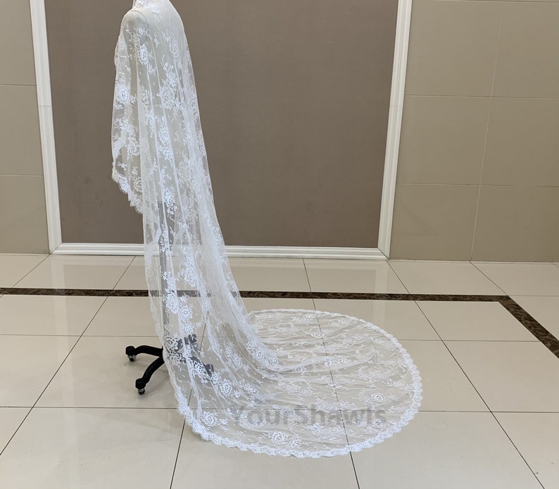 May include: A white lace wedding veil with a scalloped edge. The veil is draped over a mannequin and is flowing on the floor.