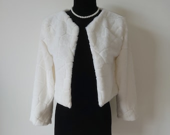 May include: A white faux fur bolero jacket with a geometric pattern. The jacket is displayed on a black mannequin and has long sleeves. A pearl necklace is visible around the mannequin's neck. The jacket is a cropped style.
