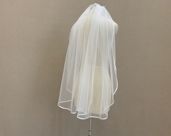 May include: A white single-tier bridal veil with a satin edge. The veil is on a white dress form.