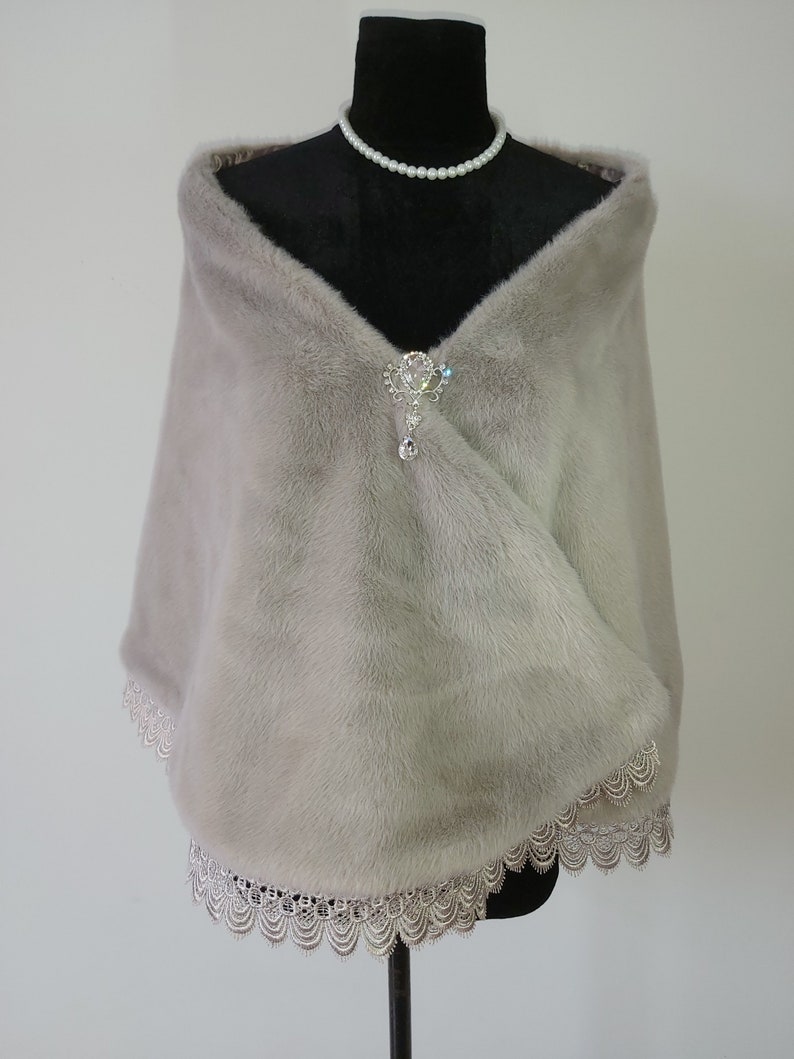 May include: A light gray faux fur shawl with a delicate lace trim. The shawl is draped over a black mannequin and features a silver and crystal brooch.