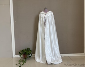 May include: A white satin hooded cape with lace trim, displayed on a mannequin. The cape is long and flowing, with a delicate lace border down the front and around the hood. The cape is a bridal or formal wear item.