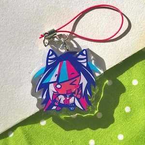 May include: A colorful anime-style charm featuring a character with blue, pink, and white hair. The charm has a red lanyard attached. The character is smiling and giving a peace sign. The background is a green dotted fabric.