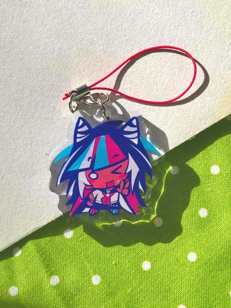 May include: A colorful anime-style charm featuring a character with blue, pink, and white hair. The charm has a red lanyard attached. The character is smiling and giving a peace sign. The background is a green dotted fabric.