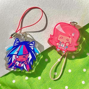 May include: Two acrylic charms featuring anime-style characters. One charm has blue, pink, and white hair, and the other has pink hair. Each charm has a silver clasp and a colored cord for attachment. The background is a green fabric with white polka dots.