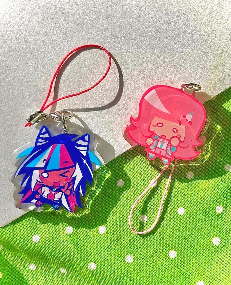May include: Two acrylic charms featuring anime-style characters. One charm has blue, pink, and white hair, and the other has pink hair. Each charm has a silver clasp and a colored cord for attachment. The background is a green fabric with white polka dots.